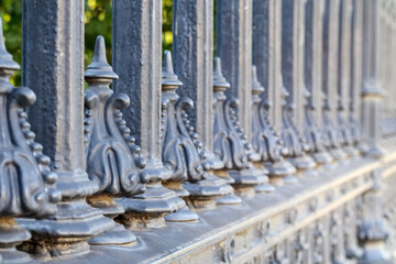 Obraz premium Decorative, dark metal fence with pointed surfaces and ornamented elements, with green trees and bushes in a blurred background.