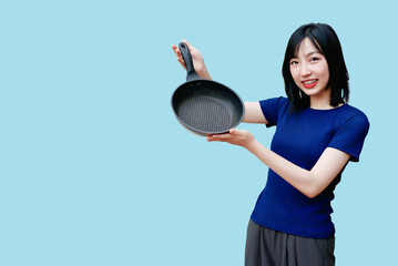 Asian woman smiles while holding a frying pan against a bright blue background, showcasing her...
