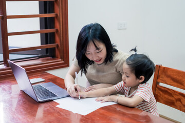 Mother teaches young child writing skills at home workstation during daytime in a cozy, well-lit...