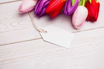 Flat design is showing tulip blossoms resting on pale wooden tabletop with blank white gift tag