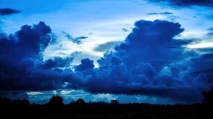 Dramatic sky with billowing deep blue-purple clouds during dawn or dusk, transitioning to lighter blue above, silhouetted trees and structures below, soft intense lighting, moody atmosphere, impending