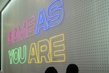 Side angle photo of a neon wall sign displaying the words "COME AS YOU ARE" in colorful lights on a perforated background inside a store.