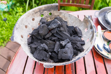 Bowl of black charcoal pieces on a wooden outdoor patio table near a chair with greenery in the background