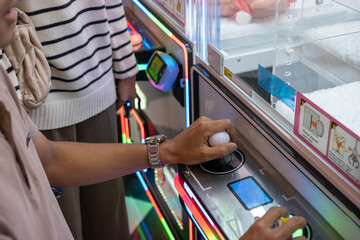 Close-up of a person's hand controlling a claw machine joystick at an arcade, surrounded by colorful LED lights and game controls.
