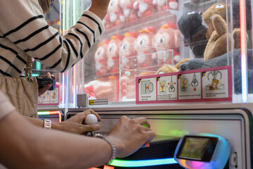 Two people using a joystick and buttons to play a colorful claw machine, surrounded by plush toys. The game area is bright and vibrant, capturing a fun and competitive arcade moment.