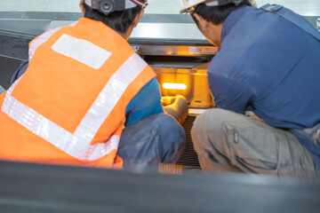 Two male workers in safety helmets and gear perform escalator maintenance, inspecting the illuminated interior section of the machinery with focused teamwork and precision.