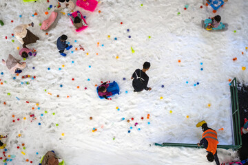Top-down view of children playing on artificial snow with colorful balls and sleds in an indoor winter-themed playground, creating a cheerful family-friendly scene.