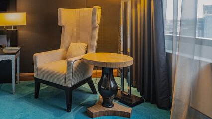 The interior of the living room. A soft armchair with a cushion in the corner. A round coffee table on one leg next to it. The lamp is lit. There are tulle curtains on the window. Blue carpet 