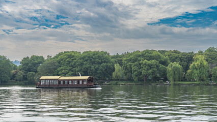 A beautiful Chinese wooden tourist boat with a tiled roof floats on a calm lake. Glare, ripples on the water. Green vegetation on the shore. Blue sky, clouds. China. Hangzhou. West Lake Xi Hu
