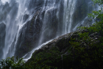 A powerful waterfall cascades over moss-covered rocks, creating a misty spray in the lush tropical forest. The dynamic flow of water contrasts beautifully with the deep green of surrounding foliage.