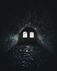 Moody interior of a dark, ancient stone chamber illuminated by soft natural light through two small...