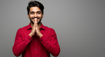 Cheerful young Indian man in red shirt standing against gray background with hands together and crossed fingers, smiling confidently with optimism and hope