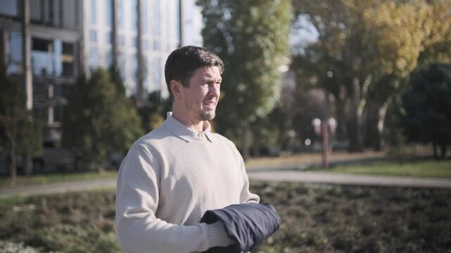 Man outdoors in city park takes off jacket, squinting due to bright sunlight