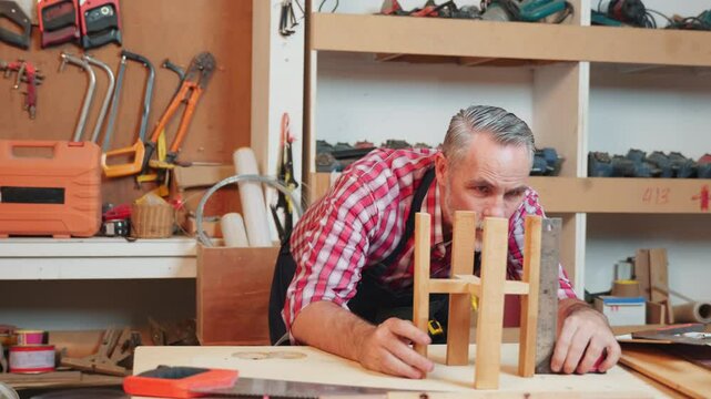 Senior carpenter uses a ruler and set square to measure and assemble a wooden structure with precise alignment, focusing on craftsmanship. Workshop is organized with tools and materials