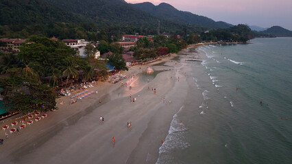 Tourists are walking on a beach in front of hotels and restaurants at sunset, with ocean waves gently lapping the shore, in a beautiful tropical paradise in thailand