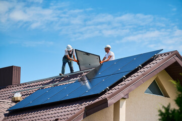 Workers building solar panel system on roof of house. Two men installers in helmets carrying...