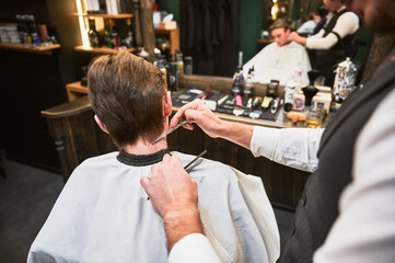 Close-up of experienced barber using comb and scissors to cut extra hair. Young client first time...