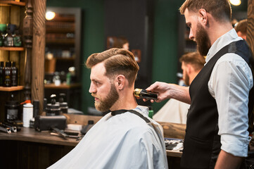 Handsome man visiting hairstylist in barbershop. Professional hairdresser using electric razor to shave nape carefully. Side view of customer getting haircut on background of big mirror.