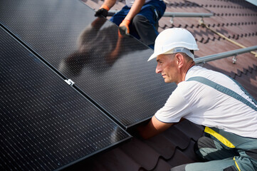 Workers building solar panel system on rooftop of house. Two men installers in helmets installing...