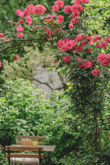 A charming rose archway frames a sun-drenched garden scene with outdoor seating.