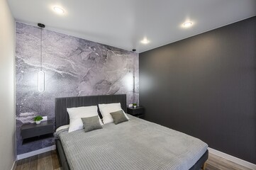modern bedroom features a grey upholstered bed with white pillows, a grey throw, and floating nightstands. Walls are grey marble and solid grey. Three ceiling lights add brightness