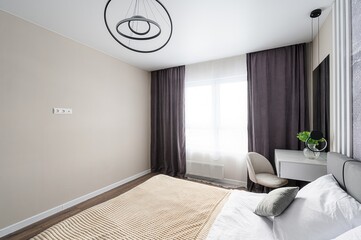 modern bedroom features neutral walls, dark curtains, a bright window, and a bed with a textured blanket. A sleek, circular light fixture hangs from the ceiling