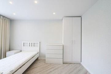 minimalist bedroom features a white bed, a small nightstand, a chest of drawers, and a wardrobe....