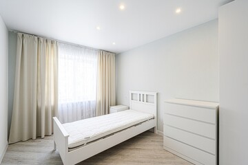 bright, minimalist bedroom features a single bed, white furniture, light beige curtains, and pale wood-look flooring. The room is clean and simple