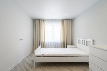 bright, minimalist bedroom with white walls and a white bed. Curtains frame a window, and a dresser stands in the corner. Clean lines and a neutral palette create a serene atmosphere