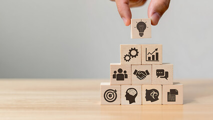 Hand placing a lightbulb block on top of a pyramid of wooden blocks with various business icons.