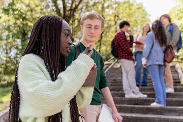 Two diverse students talking and pointing while group of classmates stands in background on sunny...