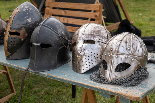 Four medieval-style helmets are displayed on a rustic table during a historical reenactment event, showcasing various armor styles.