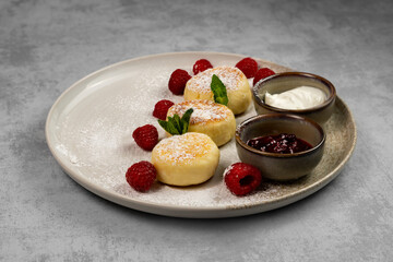Cottage cheese pancakes with powdered sugar on a plate with raspberries and jam with sour cream on a gray background. Concept of a nutritious and healthy breakfast. Top view, copy space.