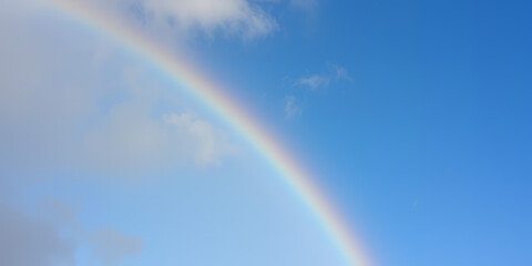 A vibrant rainbow arching across a clear blue sky with scattered soft white clouds visible
