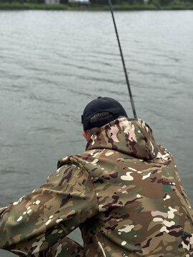 Back view of a fisherman in a multicam camouflage jacket holding a fishing rod on the river.