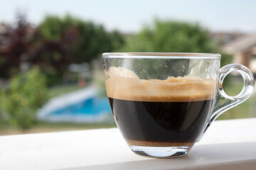 Glass cup of espresso coffee resting on white surface, with garden and pool in background