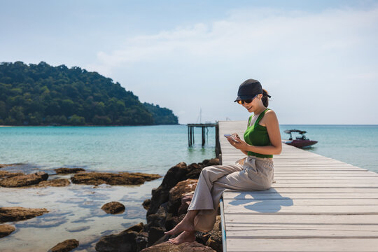 Traveler asian woman with cap relax and use mobile phone on wooden bridge on sea Bang Bao beach Koh Kood Trat Thailand Summer travel holiday vacation concept