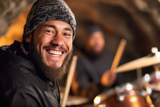 A bearded man wearing a patterned beanie smiles brightly while playing drums in a cozy, dimly lit setting.