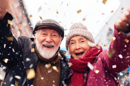 Joyful elderly couple celebrates outdoors in winter, surrounded by falling confetti and snow, expressing happiness and togetherness.