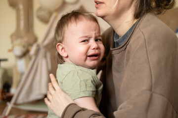 Portrait of a crying baby boy. The child is teething, he is naughty and asks for his mother hands. Mom is calming the baby. 