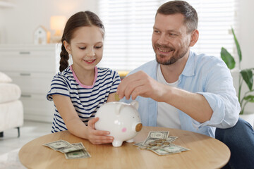 Father and daughter putting pocket money into piggybank at table indoors