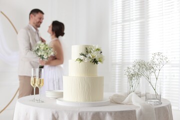 Happy married couple together indoors, focus on wedding cake and glasses of champagne on table