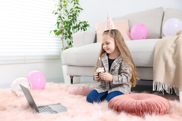 Birthday celebration. Little girl with cupcake having video call via laptop at home