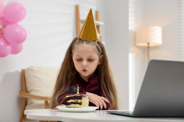 Birthday celebration. Little girl with piece of cake having video call via laptop at table indoors