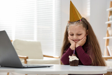 Birthday celebration. Little girl with cupcake having video call via laptop at table indoors