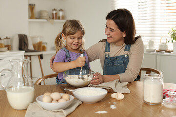 Little girl helping her mom making dough at table in kitchen