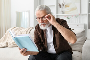 Senior man with vision problem reading book on sofa at home