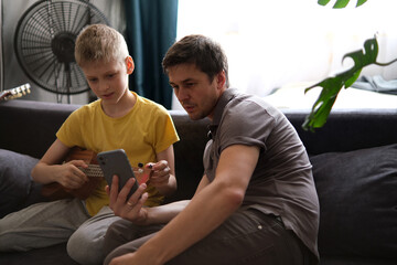 Father using smartphone teaching son to play ukulele at home