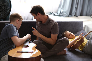 Father teaching sons guitar, relaxing together on the sofa at home