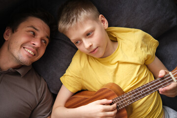 Happy father watching smiling son playing ukulele at home, relaxing on sofa together